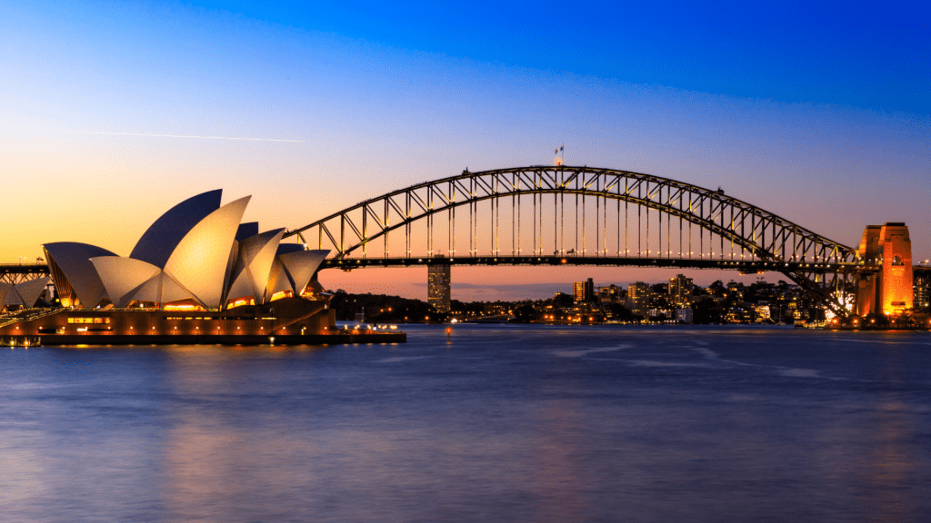 A mesmerising sunset over Sydney Harbour, with the iconic Sydney Opera House and Harbour Bridge silhouetted against a sky awash with vibrant hues of violet and gold. The radiant colours reflect upon the tranquil waters, creating a picturesque setting for the Lysosomal Disease Summit, highlighting global unity and advancement in rare disease research.